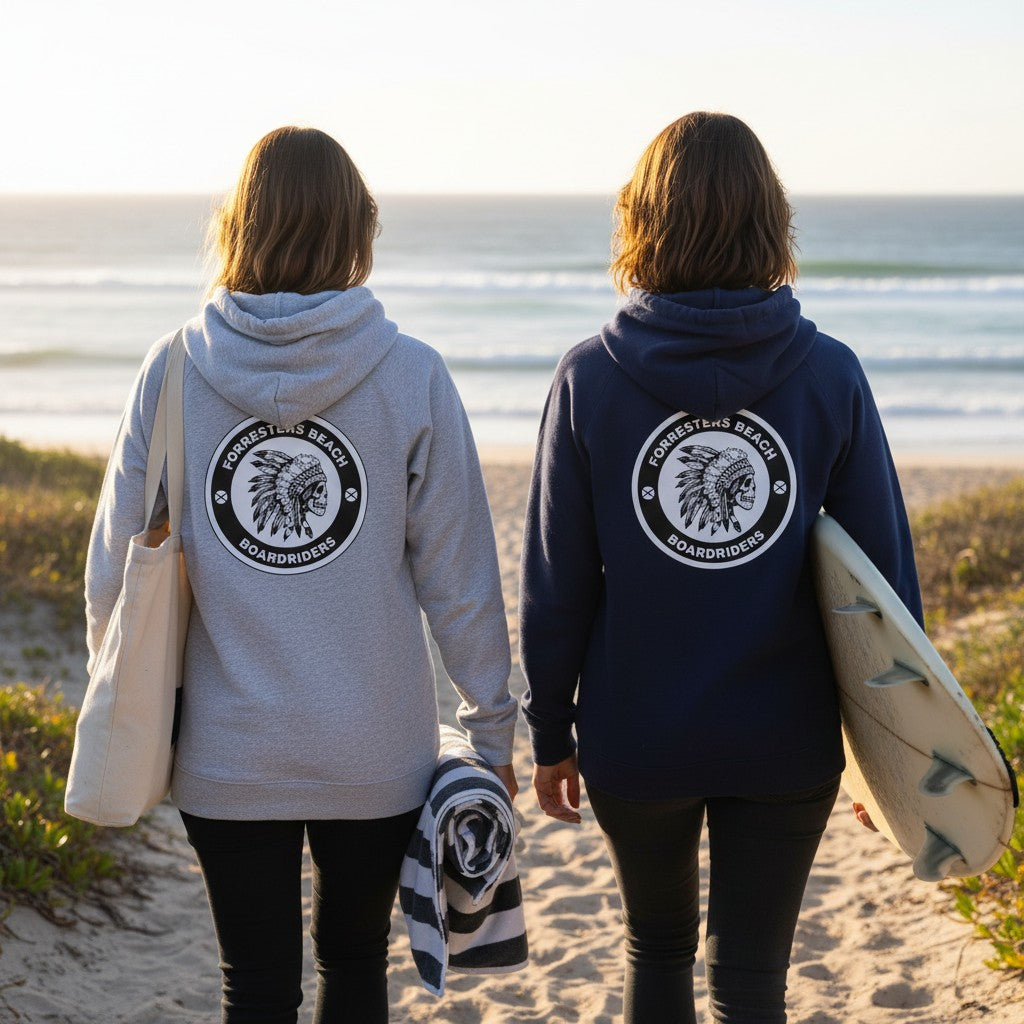 Two women wearing Sideliners Forresters Beach Boardriders hoodies in grey and navy on a beach with surfboards