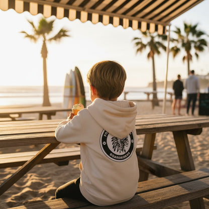 Sideliners kid wearing a cream pullover hoodie with a circular Forresters Beach Boardriders graphic on the back