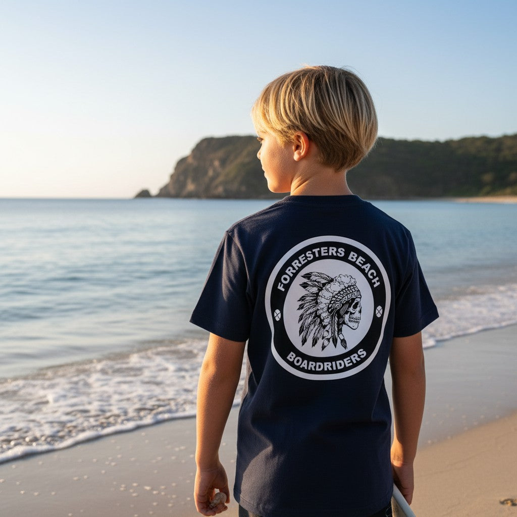 Sideliners youth navy cotton t-shirt with Forresters Beach Boardriders skull graphic on back, worn by boy at beach.